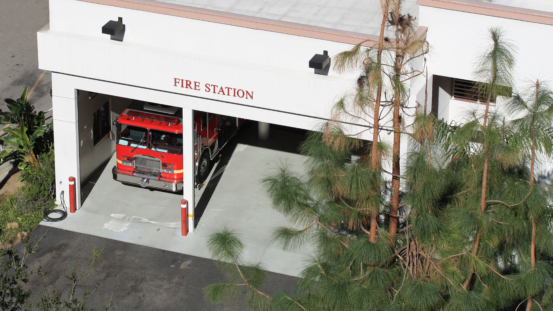 Red fire truck parked inside white fire station with pine trees nearby