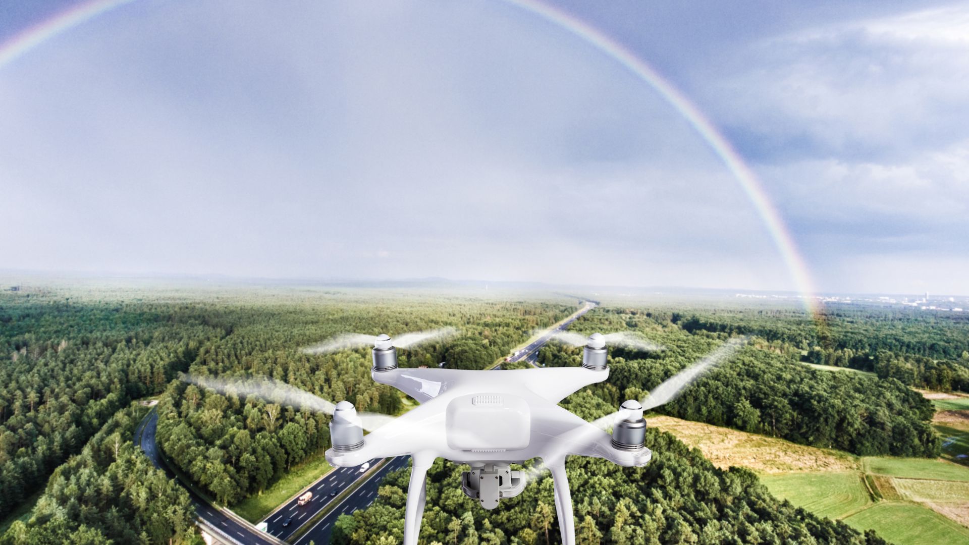 White drone flying over forest highway with rainbow in background
